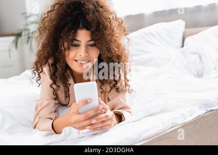 young curly woman biting lip and using smartphone in bedroom Stock Photo