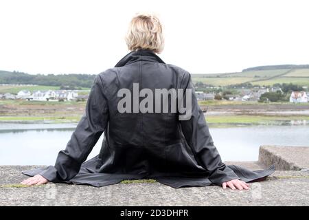 White Caucasian dyed blonde middle aged woman in her 40s wearing a leather coat jacket purchased from a second hand shop walking and sitting on harbour wall Stock Photo