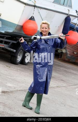 White Caucasian dyed blonde middle aged woman in her 40s wearing a blue shiny PVC raincoat purchased from a second hand shop. Pretending to dance in her raincoat and wellington with an umbrella Stock Photo