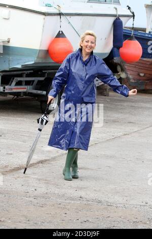 White Caucasian dyed blonde middle aged woman in her 40s wearing a blue shiny PVC raincoat purchased from a second hand shop. Pretending to dance in her raincoat and wellington with an umbrella Stock Photo