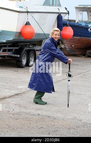 White Caucasian dyed blonde middle aged woman in her 40s wearing a blue shiny PVC raincoat purchased from a second hand shop. Pretending to dance in her raincoat and wellington with an umbrella Stock Photo