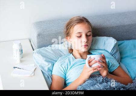 Sick teenage little girl with high fever and headache staying in bed and holding hot cup of tea. Stay at home during corona virus epidemic if you feel Stock Photo