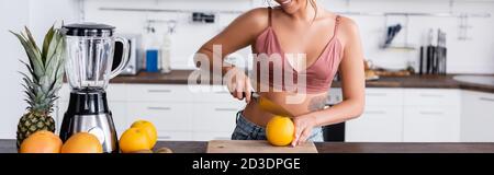 Panoramic crop of young woman cutting orange near blender in kitchen Stock Photo