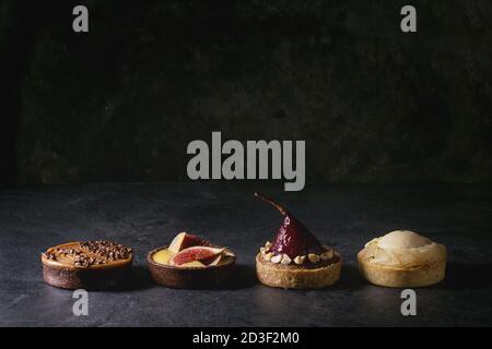 Variety of sweet tartlets with chocolate, caramel, pears, figs in row on black texture table. Stock Photo