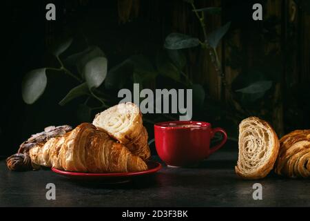 Fresh baked whole and sliced croissant with red cup of coffee espresso on black table with eucalyptus branch. Dark rustic style Stock Photo