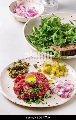 Beef tartare with quail egg in shell, cutting pickled cucumbers, capers, red onion, chives and arugula salad served in spotted ceramic plates with pep Stock Photo