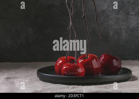 Red caramel apples sweet autumn or Christmas dessert served with branches in black ceramic plate on linen table cloth with grey wall at background. Da Stock Photo