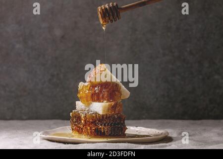 Organic honey in honeycombs and flowing honey from wooden dipper to slices of wheat bread in spotted ceramic plate on linen table cloth with grey wall Stock Photo