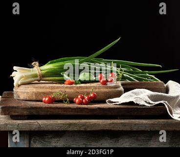 red ripe cherry tomatoes and a bundle of green onions tied with a rope lies on a brown cutting board