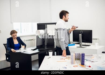 A young construction engineer is talking to someone using the speaker mode on his phone, while his colleague is doing some paperwork on the background. Stock Photo
