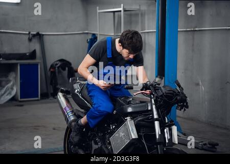 A repairer in work clothes is sitting on a bike. He uses a tablet Stock Photo