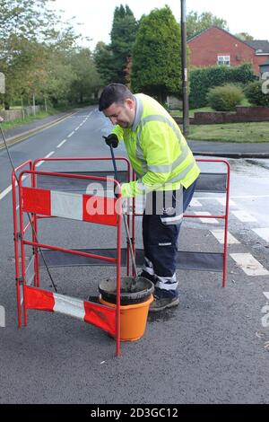 Male construction worker in the middle of the road digging a hole in the asphalt whilst wearing a hi vis jacket Stock Photo
