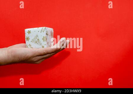 Wrapped Christmas present in a man's hand. A hand holds a small box with a gift on a red background. New Year and Christmas holidays concept Stock Photo