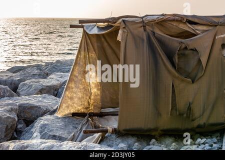 Temporary fabric tent made of old rugs standing on a stone jetty, with sunset in the background. Stock Photo