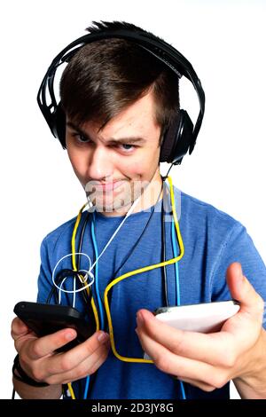 Young man with crazy expression wrapped in wires and cables ,wearing headphones and holding 2 cell phones  on white background. Stock Photo