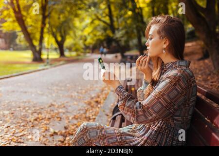 Portrait of woman applying lipstick using hand mirror in fall park sitting on bench. Girl checking makeup outdoors Stock Photo