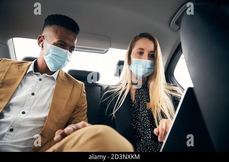 Business Couple Wearing Masks Working On Laptop In Back Of Taxi During Health Pandemic Stock Photo