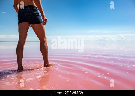 Man legs view walking in pink salt lake water with blue sky in vibrant colors. Spa recreation in summer on Syvash or Sivash, the Putrid Sea or Rotten Stock Photo