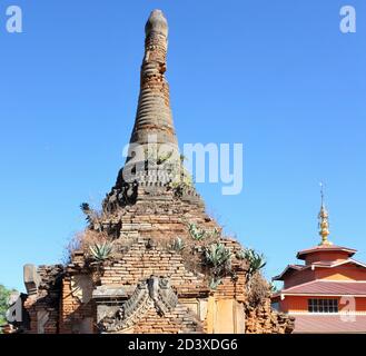 Ruins of old stone pagoda with green plants growing out of it next to a modern temple structure at In Dien located on southwestern side of Inle Lake Stock Photo