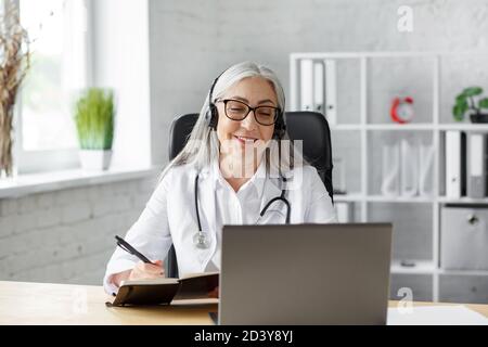 Portrait of senior grey-haired female doctor in her office using laptop for video chat with a patient. Online consultation with doctor for diagnoses Stock Photo