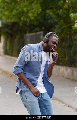 Young Afro-American man in headphones listening music on smart phone using music app. Portrait of smiling guy in earphones and mobile phone outdoors Stock Photo