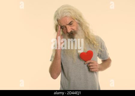 Studio shot of senior bearded man holding red heart while having headache Stock Photo