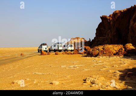 Four-wheel drive vehicles are parked next to a rocky outcrop on the salt flats of the Danakil Depression beneath a pale blue sky in Hamadella, Ethiopi Stock Photo