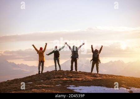 Four happy tourists stands with raised arms at mountain top against sunset mountains and sky