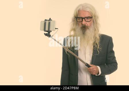Studio shot of senior bearded businessman taking selfie picture with mobile phone on selfie stick Stock Photo