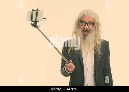 Studio shot of happy senior bearded businessman smiling while taking selfie picture with mobile phone on selfie stick Stock Photo