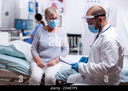 Doctor wearing face mask agasint covid-19 for safety precaution and reading test results to elderly woman in hospital. Healthcare medical physician consultation during COVID-19 global crisis. Stock Photo