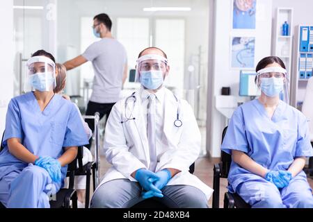 Group of medical personnel in hospital waiting area with visor and face mask against covid-19 to prevent infection. Wearing white coat and stethoscope. Stock Photo