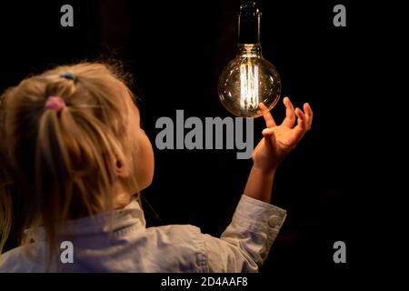 Cute adorable caucasian blond girl portrait smiling and holding in hand one of hanged edison light bulb at forest outdoor. Right solution choice Stock Photo
