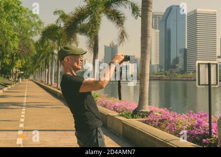 Profile view of senior tourist man wearing cap while taking pictures at peaceful park in Bangkok Thailand Stock Photo