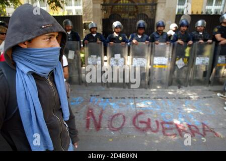 A masked protester stands outside the British embassy, protected by a line of riot police along pavement painted with anti-war slogans, in Mexico City March 20, 2003. Students outside the embassy demonstrated against the U.S.-led attack on Iraq. Graffiti on pavement reads 'No War'.
