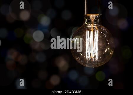 Glowing vintage old one tungsten lightbulb hanged on ceiling against black wall bokeh background. Retro styled decoration electric lamp. Christmas Stock Photo