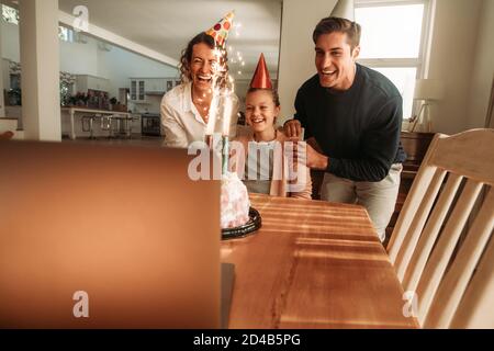 Family celebrating their daughter's birthday at home with friends over a video call on laptop. Family wearing party hats with birthday cake on table a Stock Photo