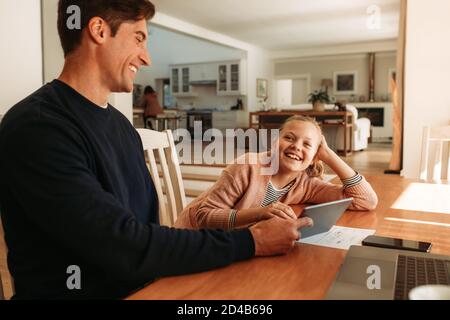 Man with his daughter sitting at table and smiling. Man holding a digital tablet and talking with his daughter at home. Stock Photo