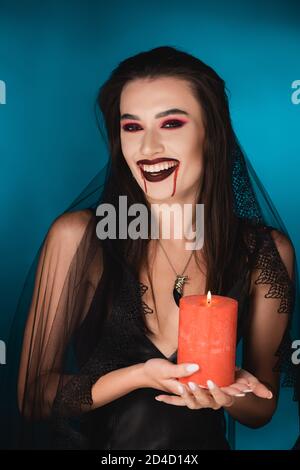 woman in veil with blood on face holding red burning candle and laughing on blue Stock Photo
