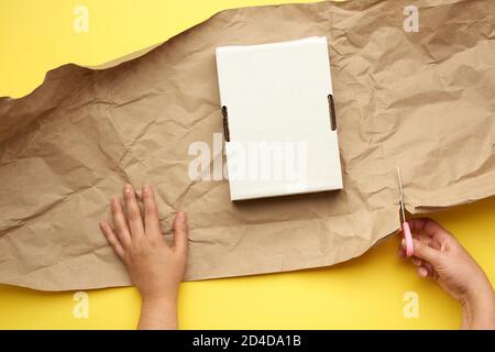 rectangular white cardboard box and two female hands are wrapped with brown paper on a yellow background, cuts off the edge with scissors Stock Photo