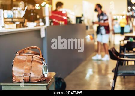 Mini portable alcohol gel bottle to kill Corona Virus(Covid-19) hang on a brown leather shoulder bag on chair in coffee shop.New normal lifestyle. Hea Stock Photo