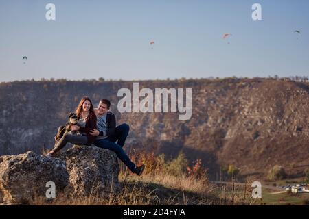 Young family with a schnauzer dog travel. Beautiful Girl hugs a young man and holds a pet in her arms. Sitting on a canyon and looking at paragliders Stock Photo