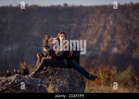 Young family with a schnauzer dog travel. Beautiful Girl hugs a young man and holds a pet in her arms. Sitting on a canyon and looking at paragliders Stock Photo