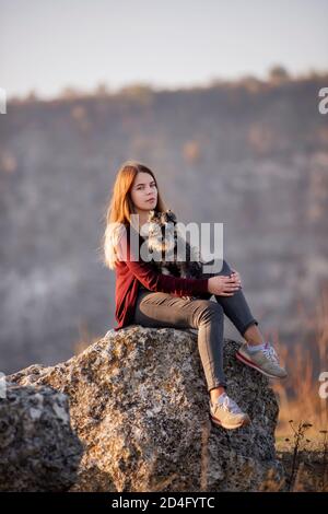 A beautiful girl with red hair is holding a Schnauzer dog in her hands, smiling and looking at the camera, sitting on a high stone. Traveling with pet Stock Photo