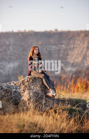 A beautiful girl with red hair is holding a Schnauzer dog in her hands, smiling and looking at the camera, sitting on a high stone. Traveling with pet Stock Photo