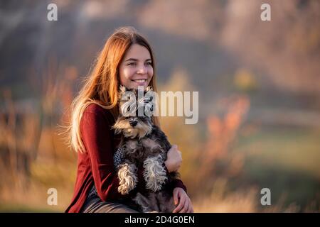 A beautiful girl with red hair is holding a Schnauzer dog in her hands, smiling and looking at the camera, sitting on a high stone. Traveling with pet Stock Photo