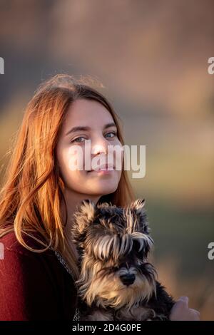 A beautiful girl with red hair is holding a Schnauzer dog in her hands, smiling and looking at the camera, sitting on a high stone. Traveling with pet Stock Photo