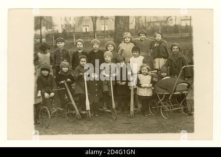 1920's postcard of group young children with wooden scooters, enjoying themselves holding scooters and a pram, outside in a park, circa 1921 U.K. Stock Photo