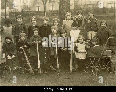 1920's postcard of group young children with wooden scooters, enjoying themselves holding scooters and a pram, outside in a park, circa 1921 U.K. Stock Photo