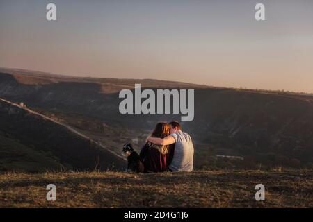 Happy couple in love are resting on the mountainside with a black Schnauzer, sitting on the ground, hugging a dog, watching bright paragliders fly in Stock Photo